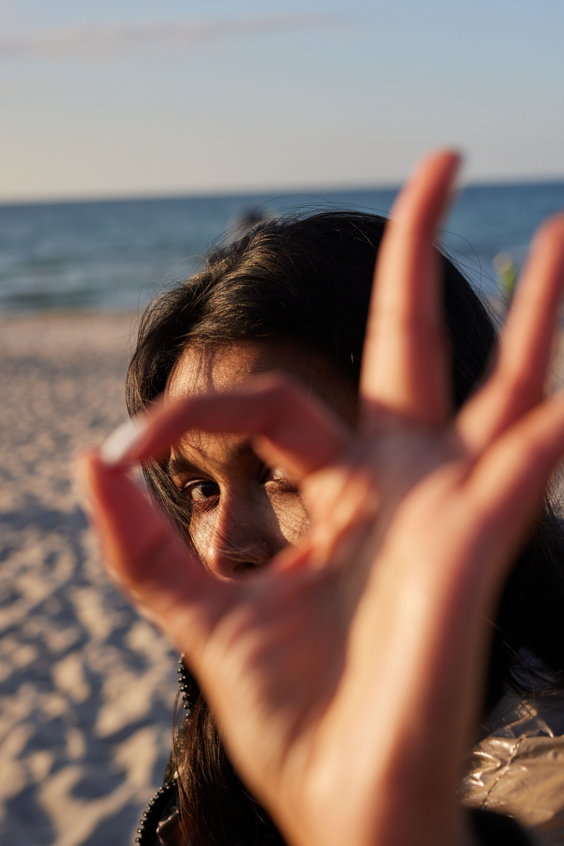 Frau macht mit ihrer Hand ein „Okay“-Zeichen, Strand und Meer im Hintergrund.