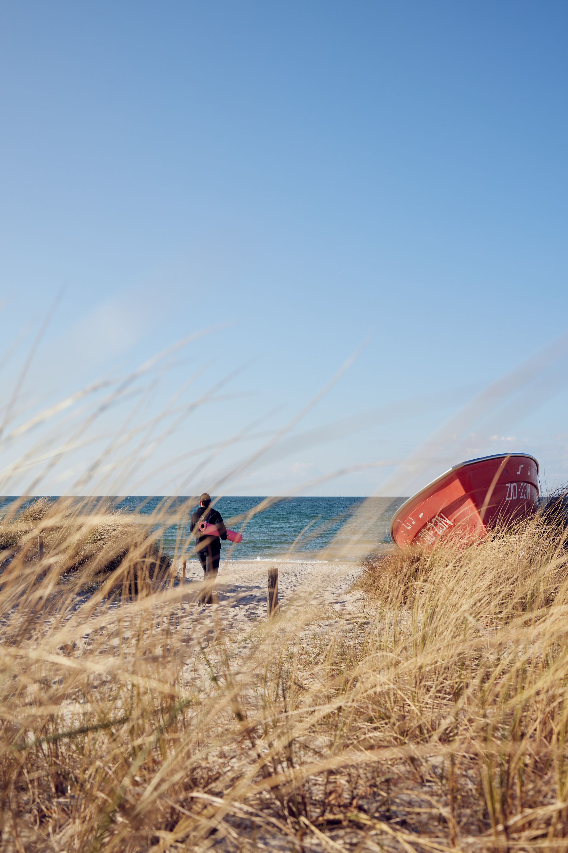 Entdecke die Vielfalt im Strandhotel Zingst Person, die am Ostseestrand mit einer roten Yogamatte spazieren geht, Dünengras im Vordergrund, blauer Himmel und die Ostsee im Hintergrund.