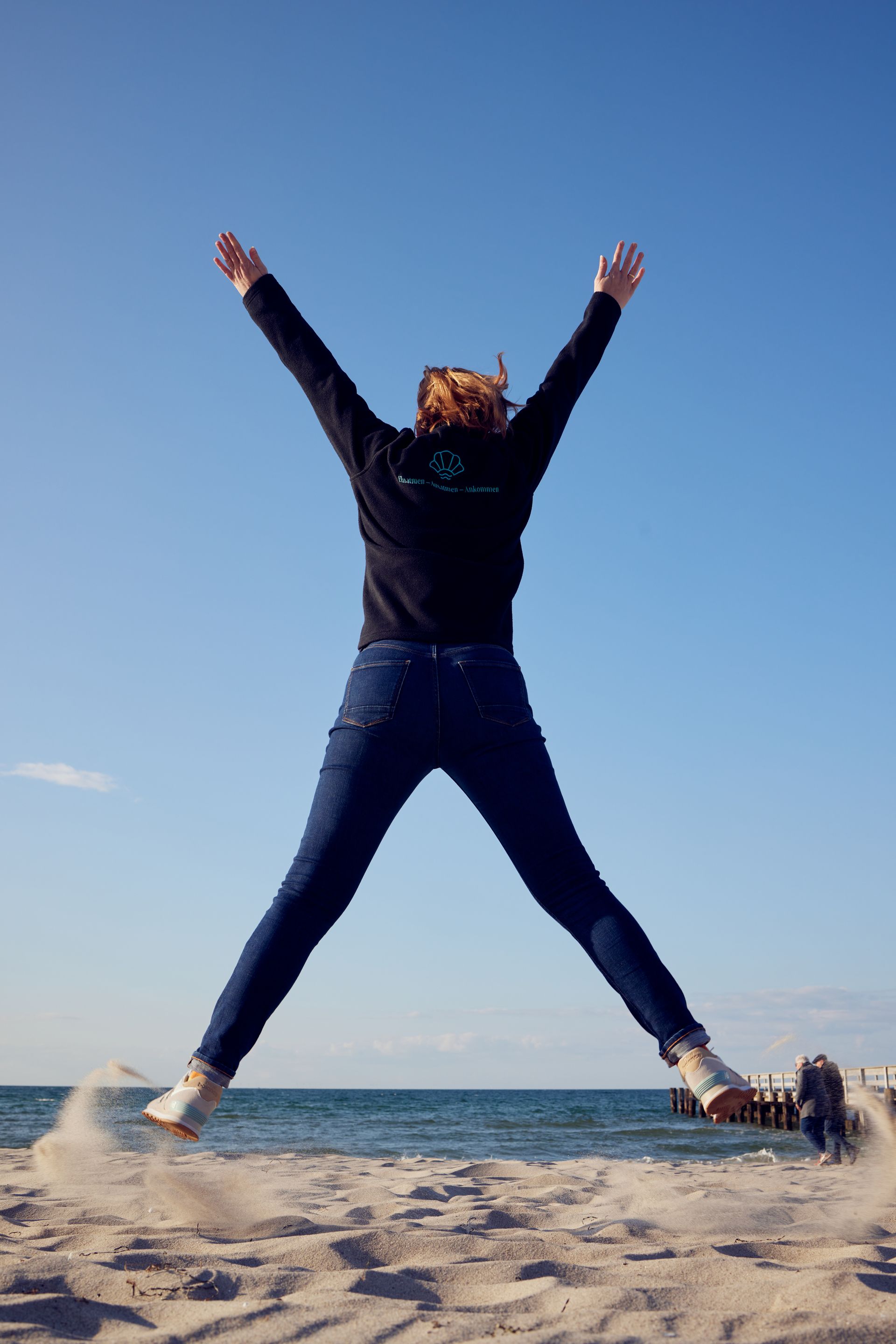 Eine Person springt mit ausgestreckten Armen und Beinen am Ostseestrand unter blauem Himmel.