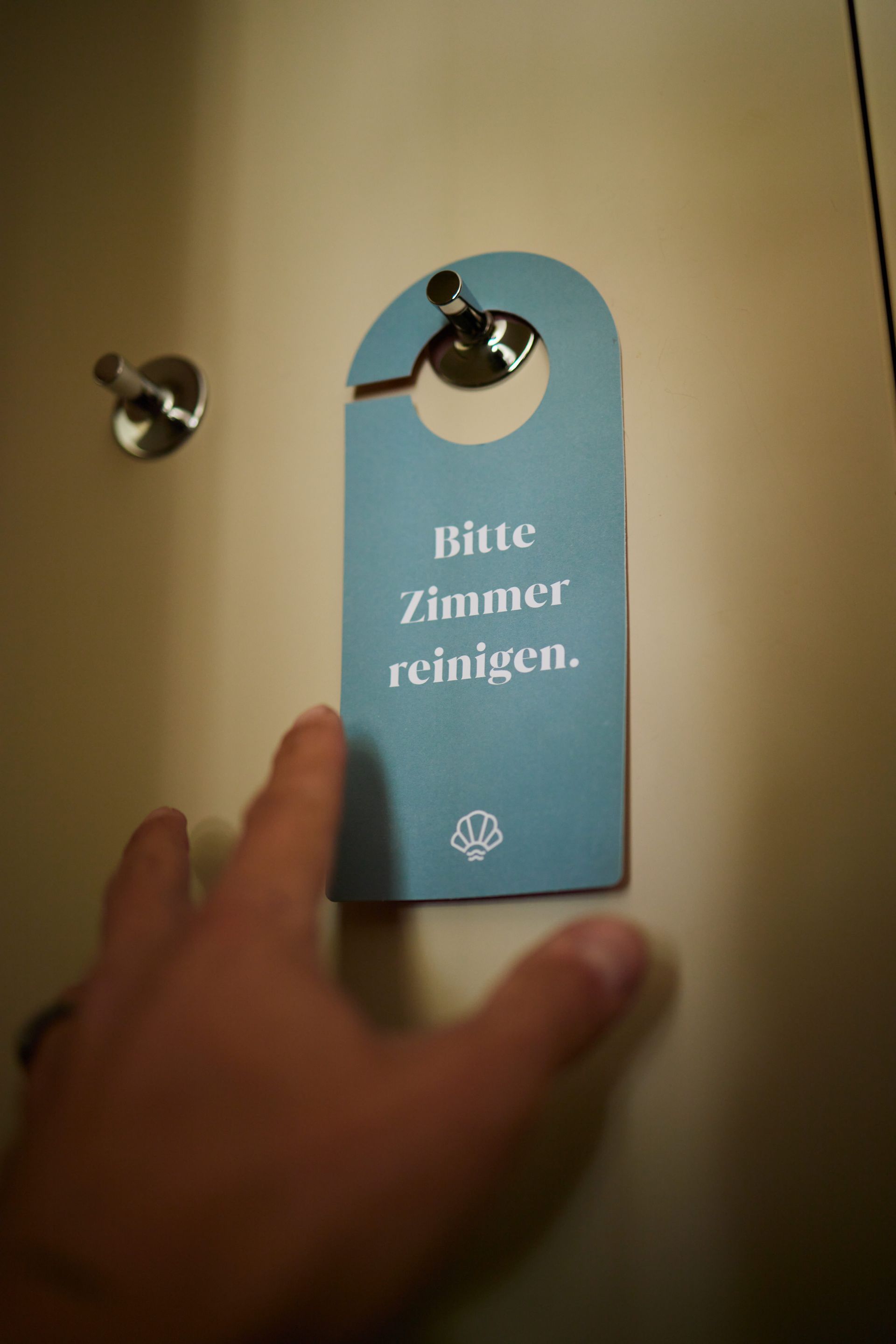 Schau hinter die Kulissen eines erstklassigen Hotels Eine Hand greift nach einem Türschild an einer beigen Tür. Auf dem Schild steht in Blau „Bitte Zimmer reinigen“.