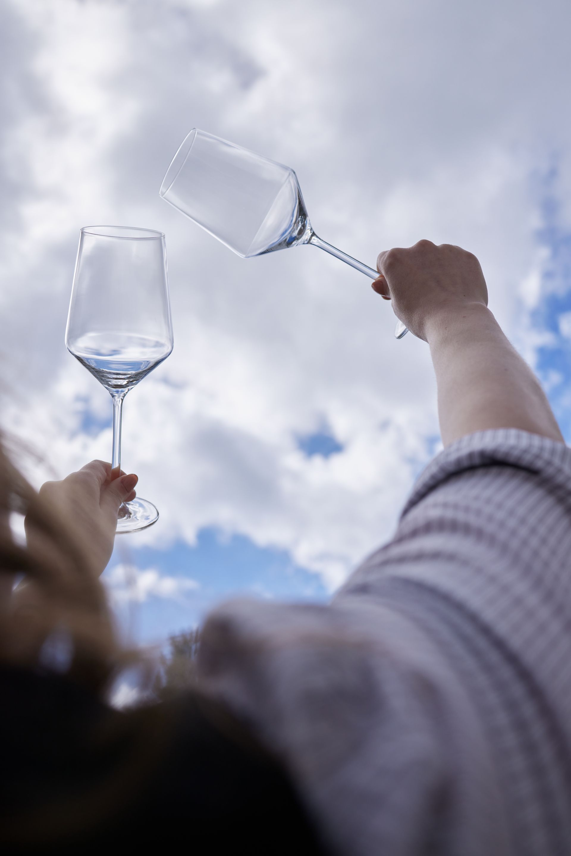 Person, die zwei leere Weingläser vor einem bewölkten blauen Himmel hält.