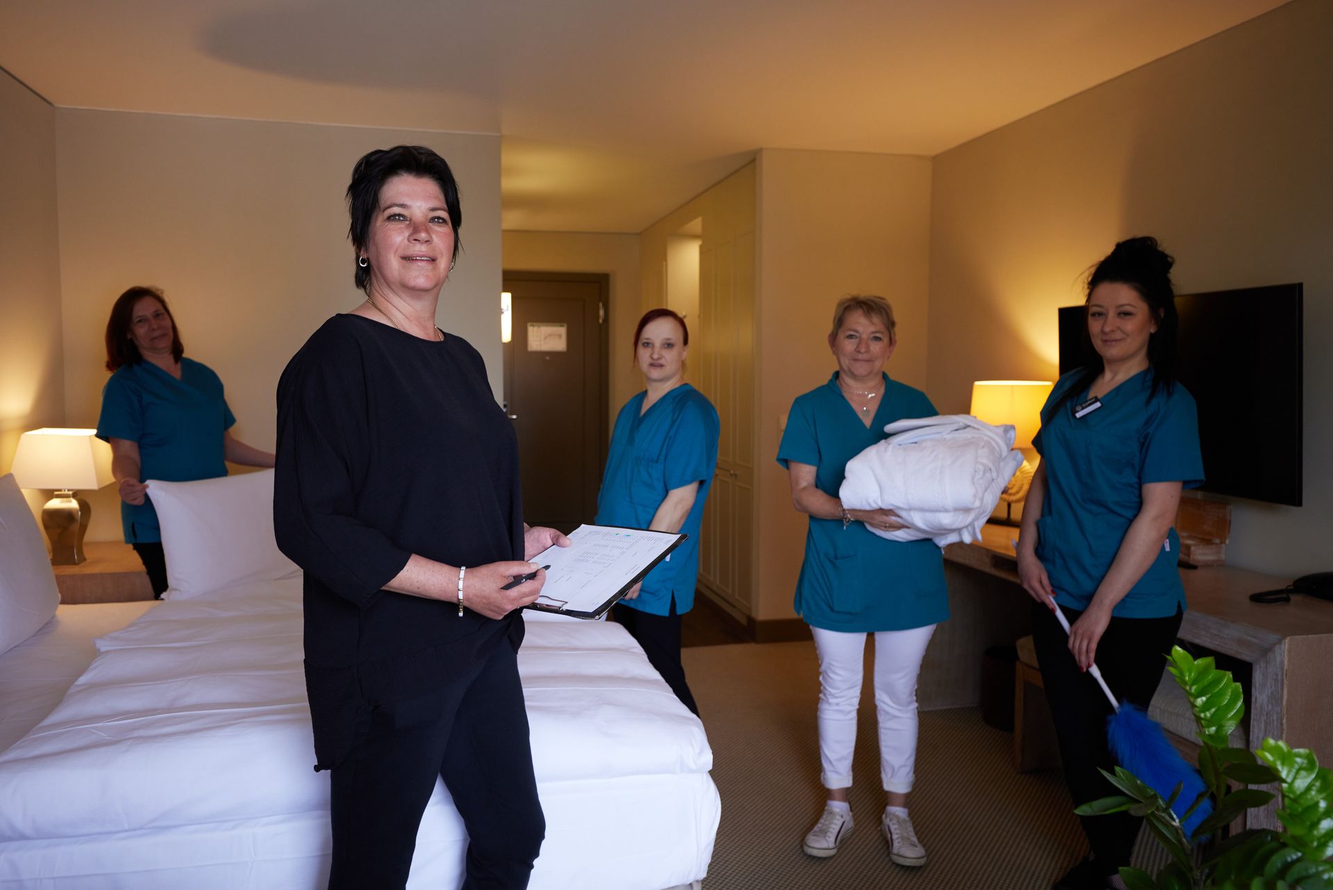 Hotel-Housekeeping-Personal in einem Zimmer, einer hält Checkliste, andere halten Bettwäsche und einen Staubwedel in der Hand.