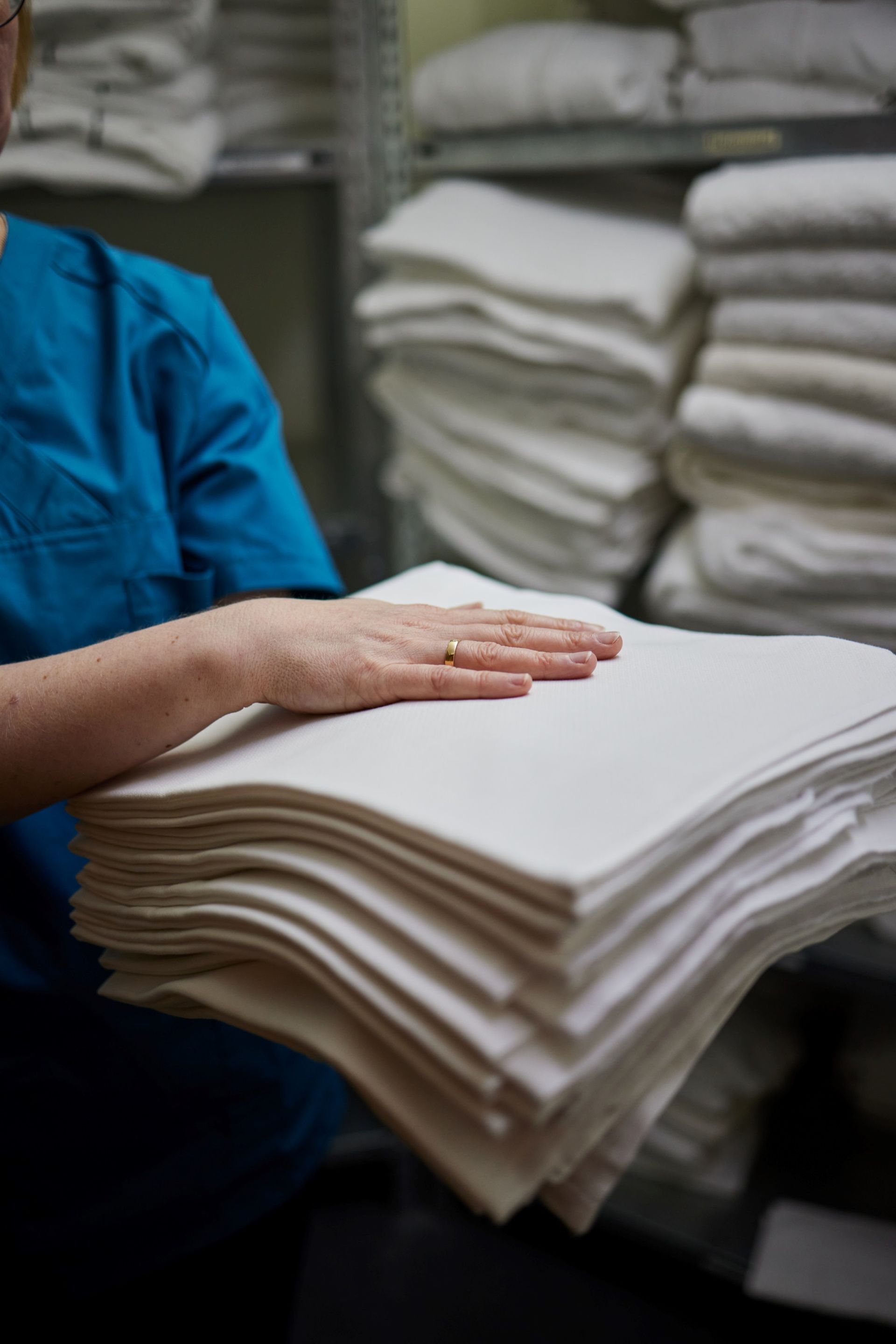 Person im blauen Kittel hält einen Stapel ordentlich gefalteter weißer Wäsche in einem Lagerraum.