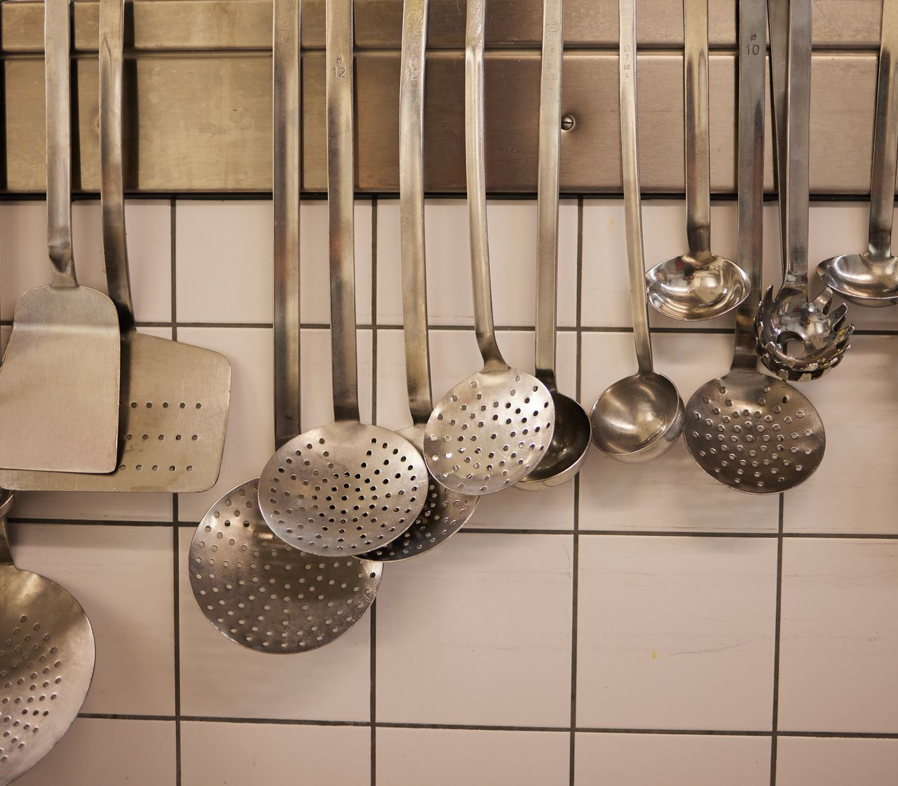 An einer gefliesten Wand hängen Küchenutensilien, darunter Pfannenwender und Schaumlöffel.