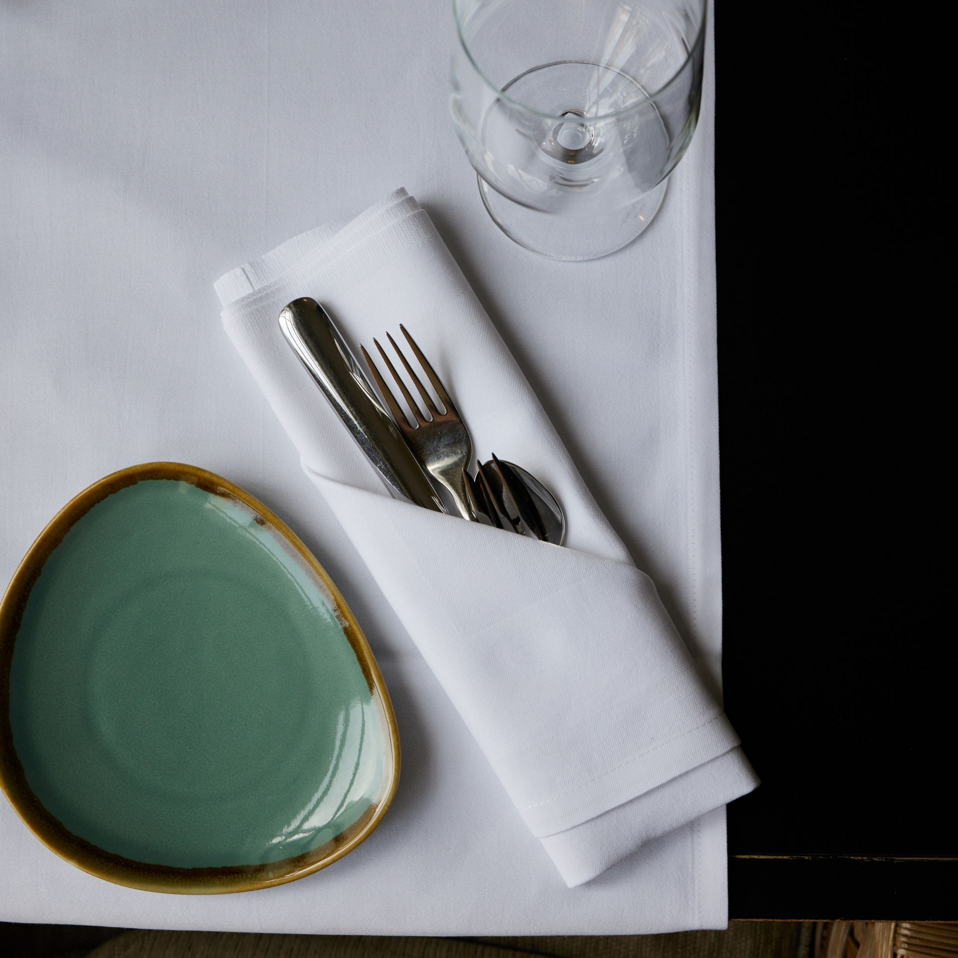 Tischgedeck mit Silberbesteck in einer gefalteten Serviette, einem blaugrünen Teller und einem Weinglas auf einer weißen Tischdecke.