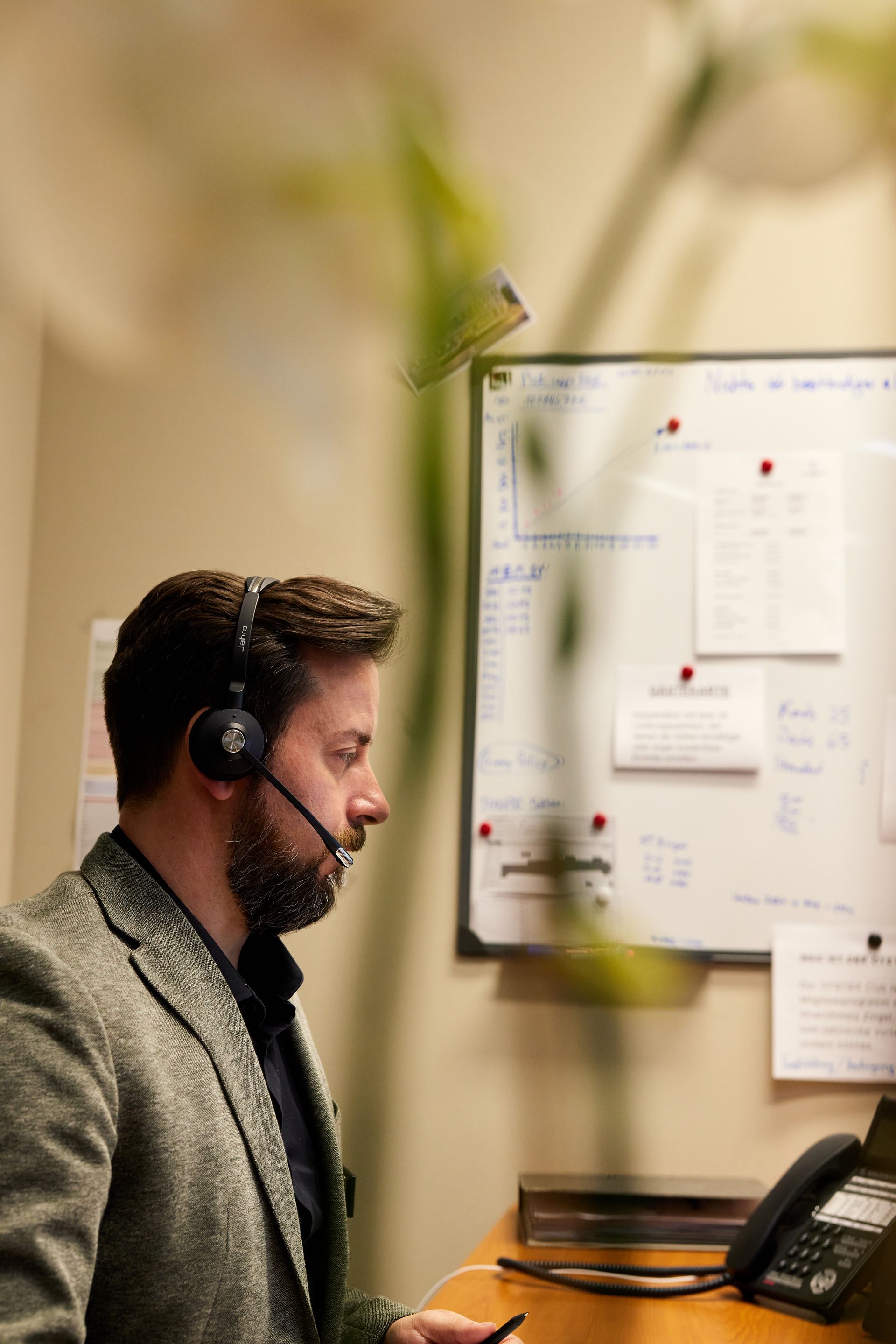 Mann mit Headset, der in einem Büro auf ein Whiteboard blickt. Im Vordergrund ist eine Pflanze zu sehen.