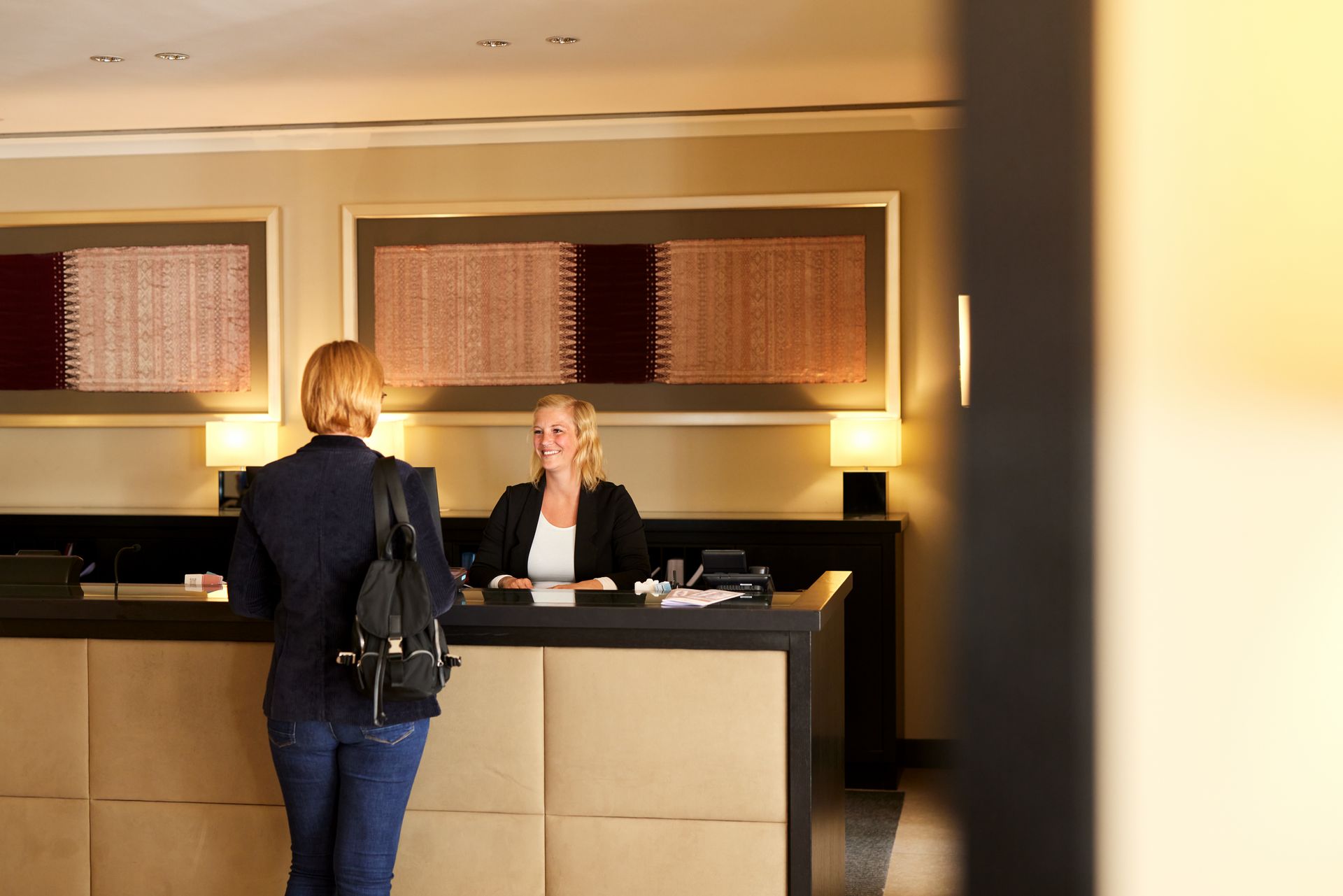 Frau an der Hotelrezeption im Gespräch mit einem Hotelangestellten. Beiger Schreibtisch, Kunstwerke und neutrale Farben.