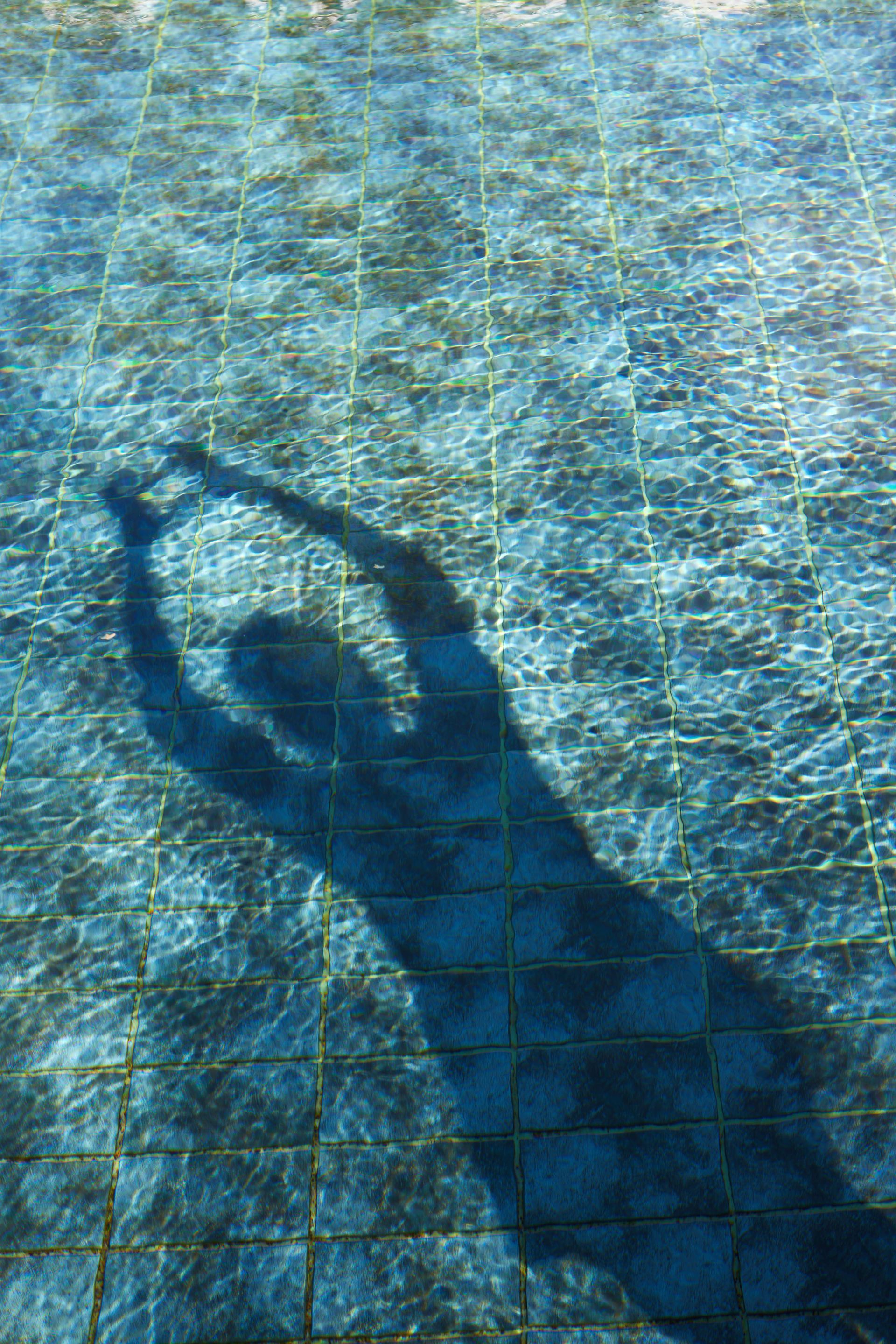 Entspannung und Wohlfühlmomente für unsere Gäste Schatten einer Person in einem Pool, Arme erhoben; blaues Wasser und gefliester Poolboden.