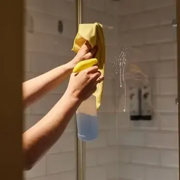 Hände besprühen und wischen eine Glasduschtür mit einem gelben Tuch und Reiniger ab.