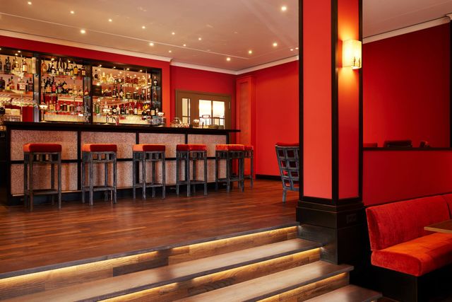 Blick in die Oyster Bar mit roten Barhockern und  hinterleuchteter Bartheke, Holzboden und einer kleinen Treppe.
