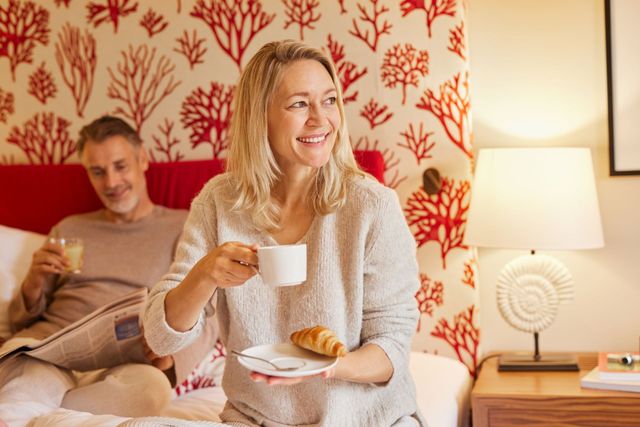 Frau lächelt mit Kaffee und Croissant im Bett, Mann hinter ihr liest Zeitung. Schlafzimmerszene.