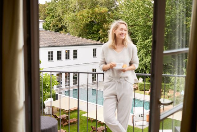 Frau auf dem Balkon lächelt, hält eine Kaffeetasse in der Hand und blickt auf einen Pool und auf das Strandhotel zingst.