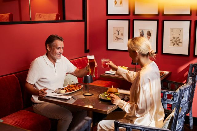 Ein Paar stößt in einem Restaurant mit Getränken an; rote Wände, gerahmte botanische Kunstwerke, ein Tisch mit Speisen.