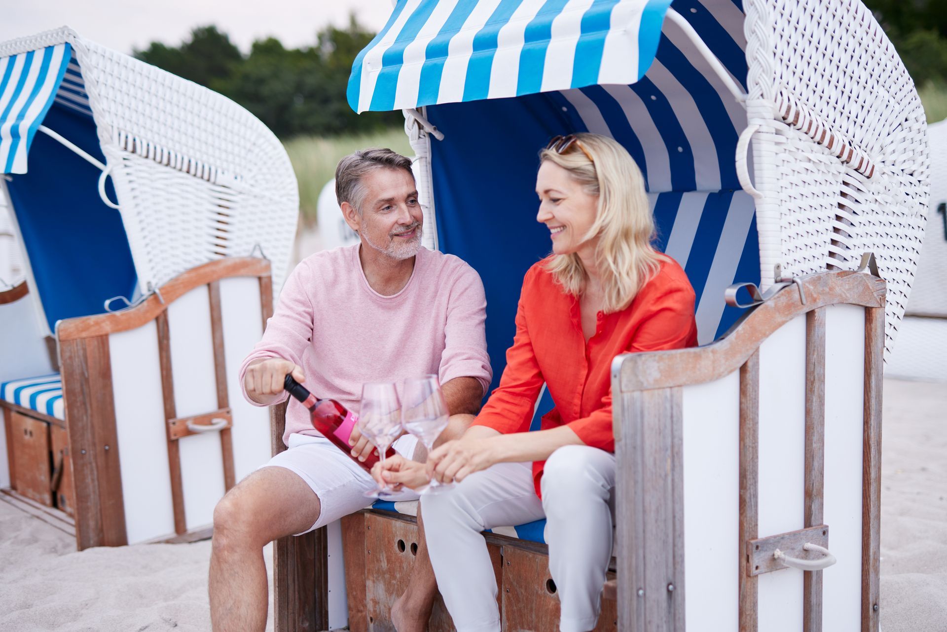 Ein Pärchen teilt sich ein Glas Wein und sitzt in Strandstühlen unter blau-weiß gestreiften Sonnensegeln.