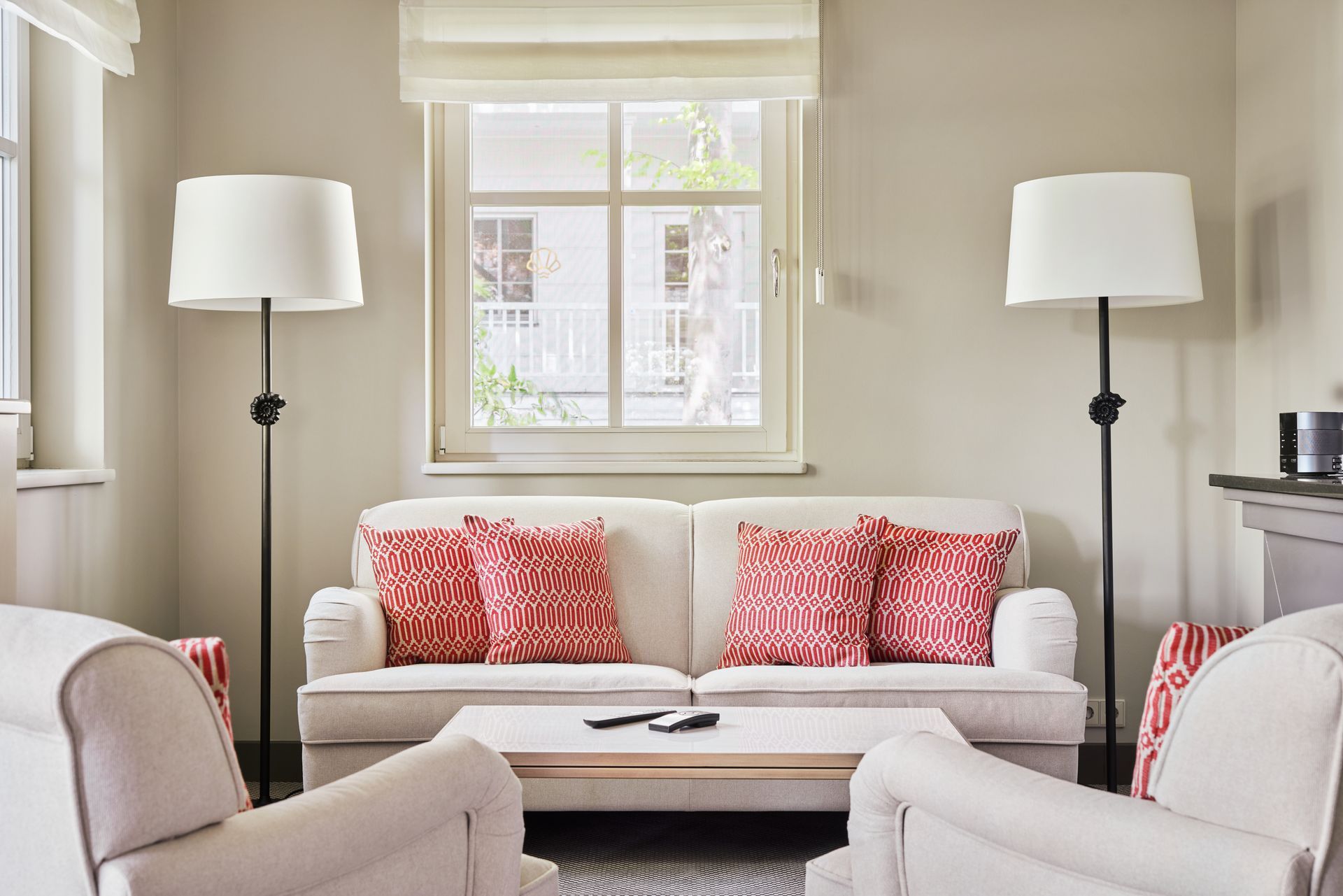 Blick ins Kapitänshaus. Wohnzimmer mit Sofa, Stühlen, Lampen und einem Fenster. Beige Wände, rote Kissen.