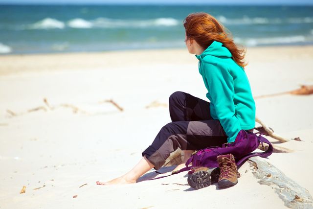 Eine Frau in einem türkisfarbenen Kapuzenpulli sitzt am Strand und blickt aufs Meer. Wanderschuhe und ein lila Rucksack.