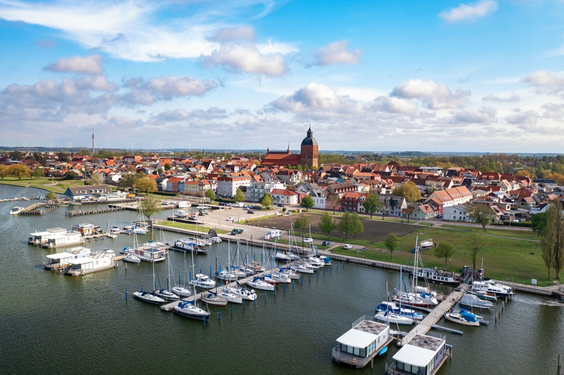 Luftaufnahme eines Hafens mit vielen Booten vor einer Stadt mit roten Dächern und einem hohen Kirchturm unter blauem Himmel.
