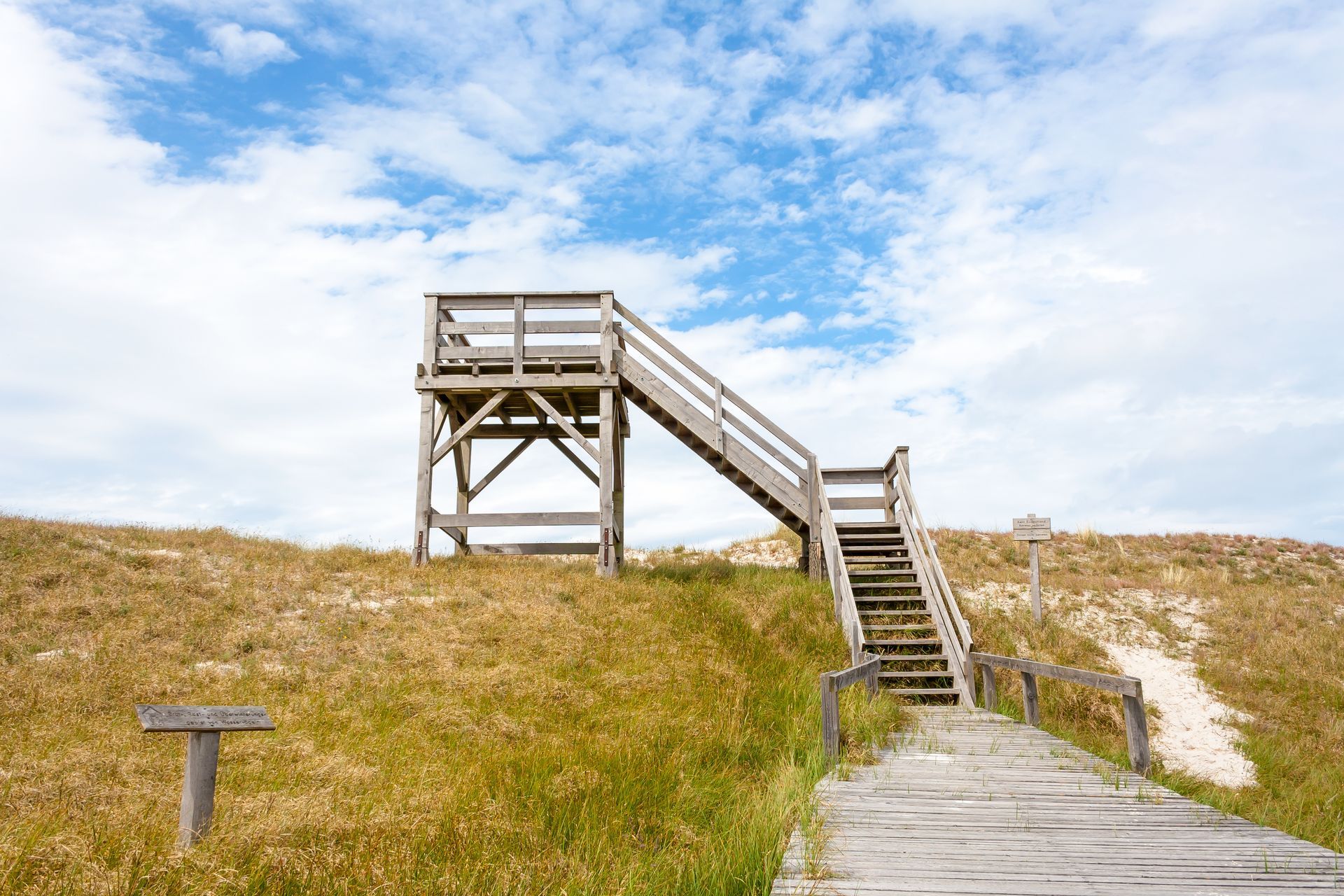 Holztreppe und Aussichtsplattform auf einer grasbewachsenen Düne unter blauem Himmel.
