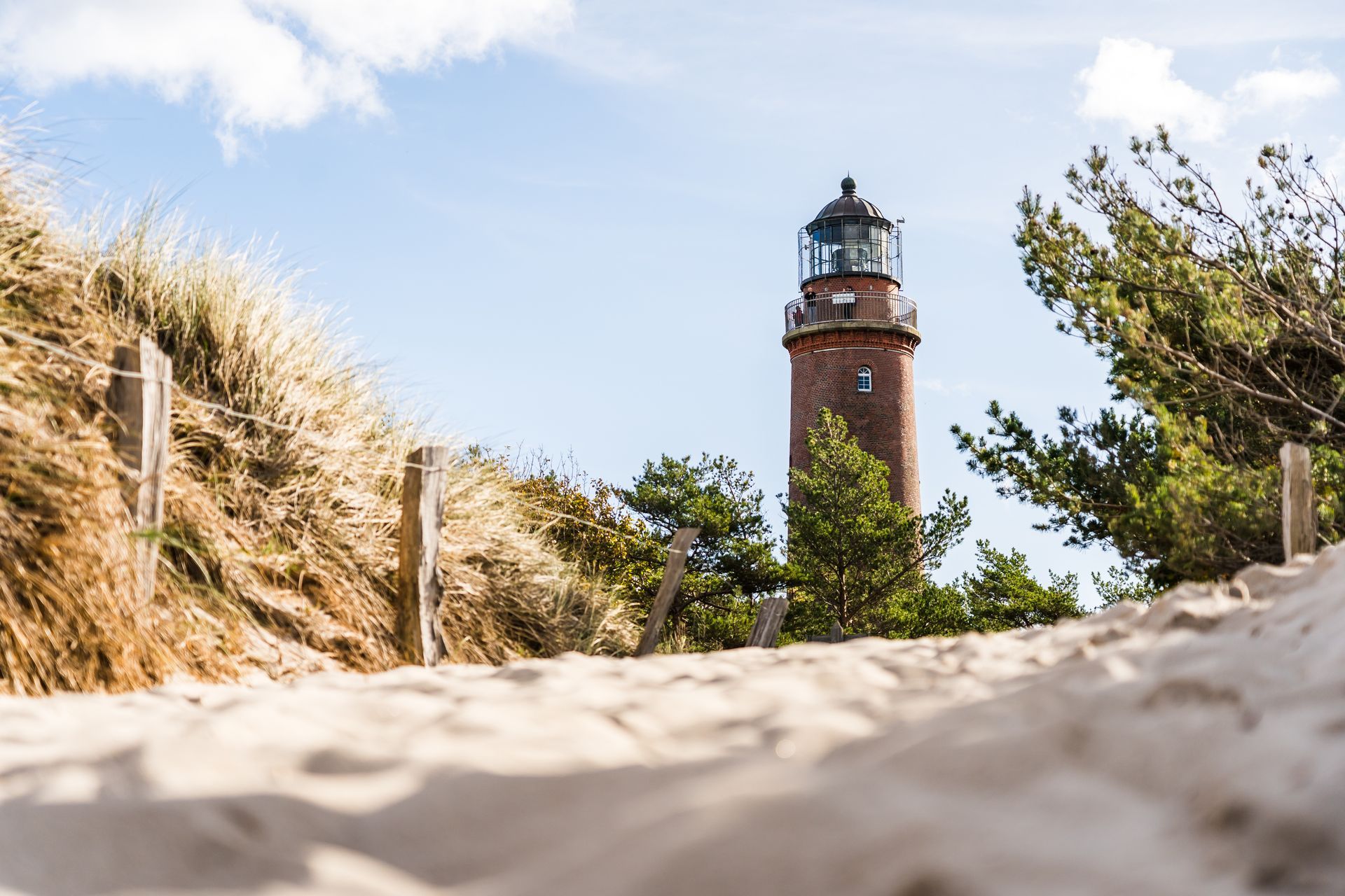 Ein sandiger Pfad führt unter blauem Himmel zu einem Leuchtturm aus Backstein, umgeben von Dünengras und Bäumen.
