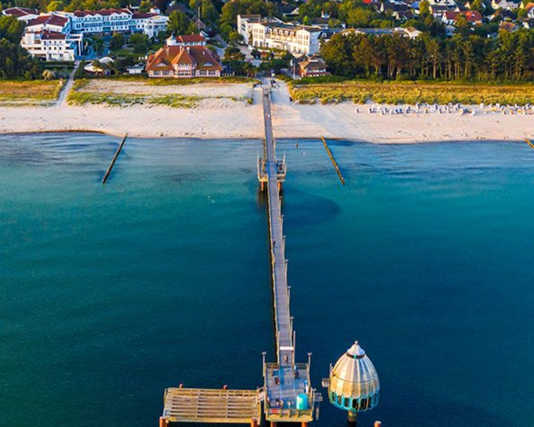 Ein hölzerner Pier erstreckt sich ins türkisfarbene Meer; im Hintergrund sind ein Sandstrand und Gebäude zu sehen.