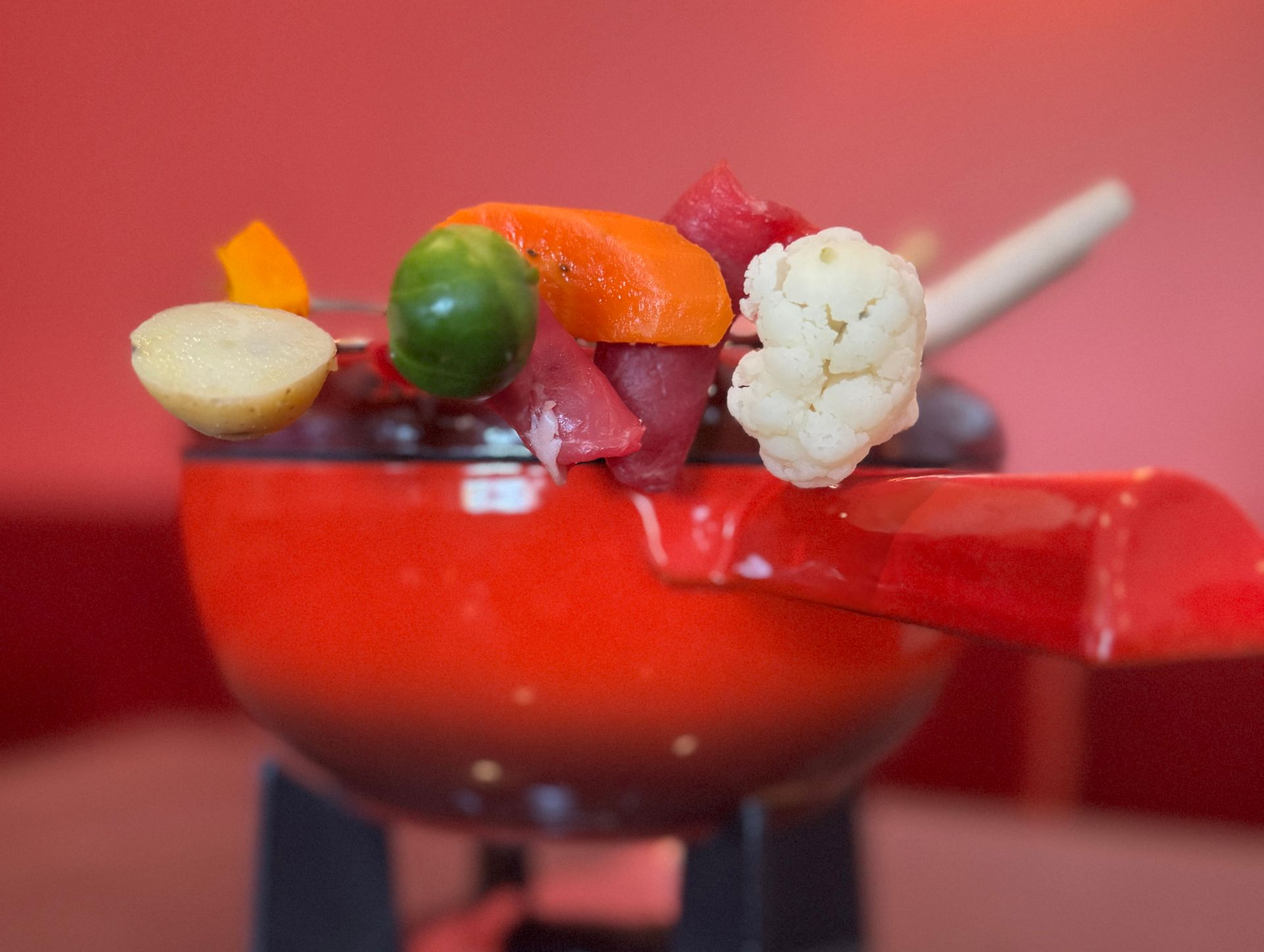 Drei Lebensmittel auf Fonduegabeln: grün, gelb und rot vor rosa Hintergrund.