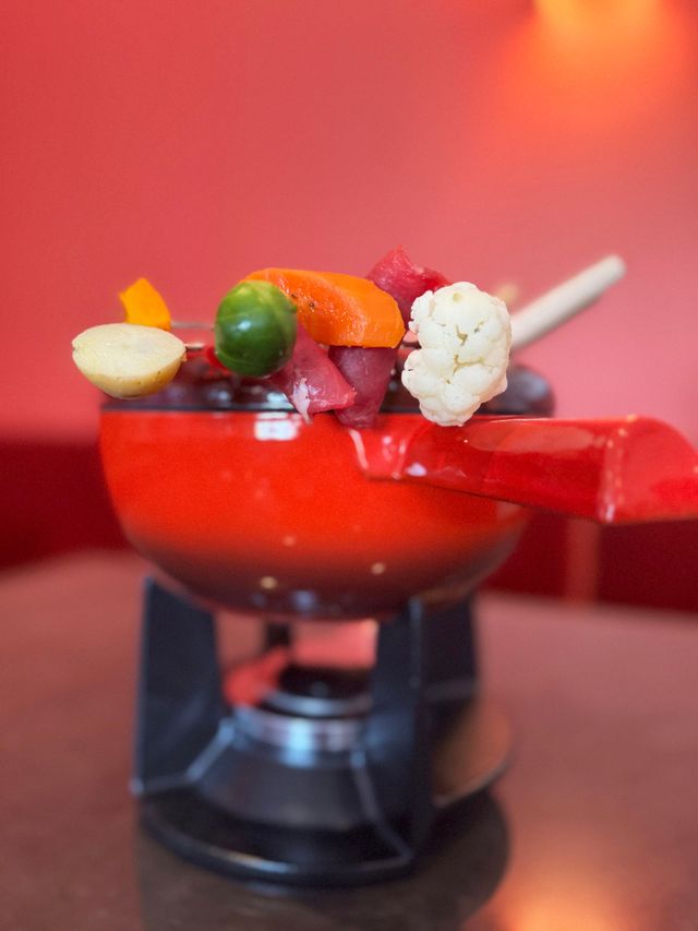 Roter Fonduetopf mit verschiedenen Gemüsesorten vor rotem Hintergrund.