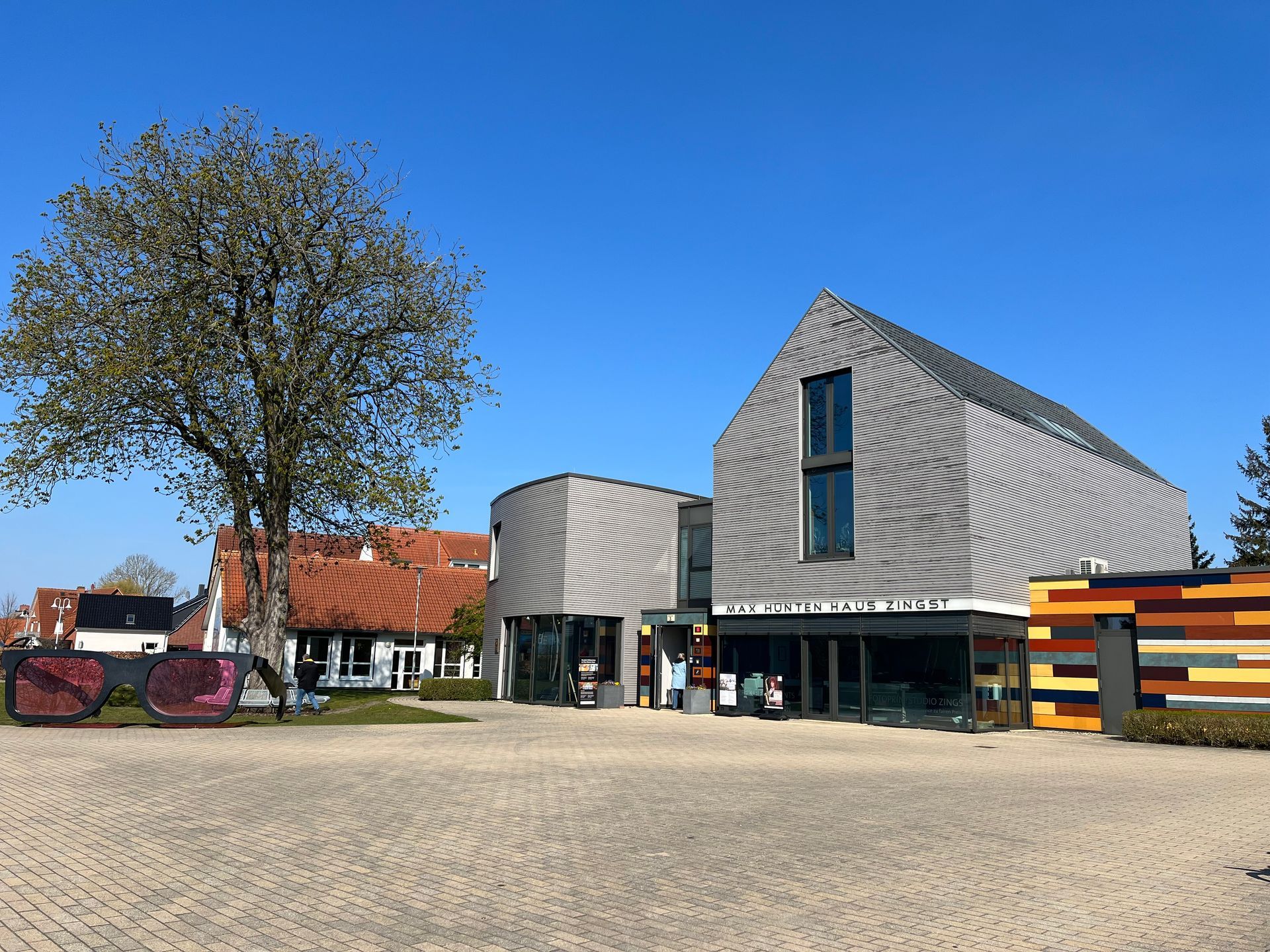 Moderner Gebäudekomplex mit gestreifter Fassade, Baum und Kopfsteinpflasterhof an einem sonnigen Tag.