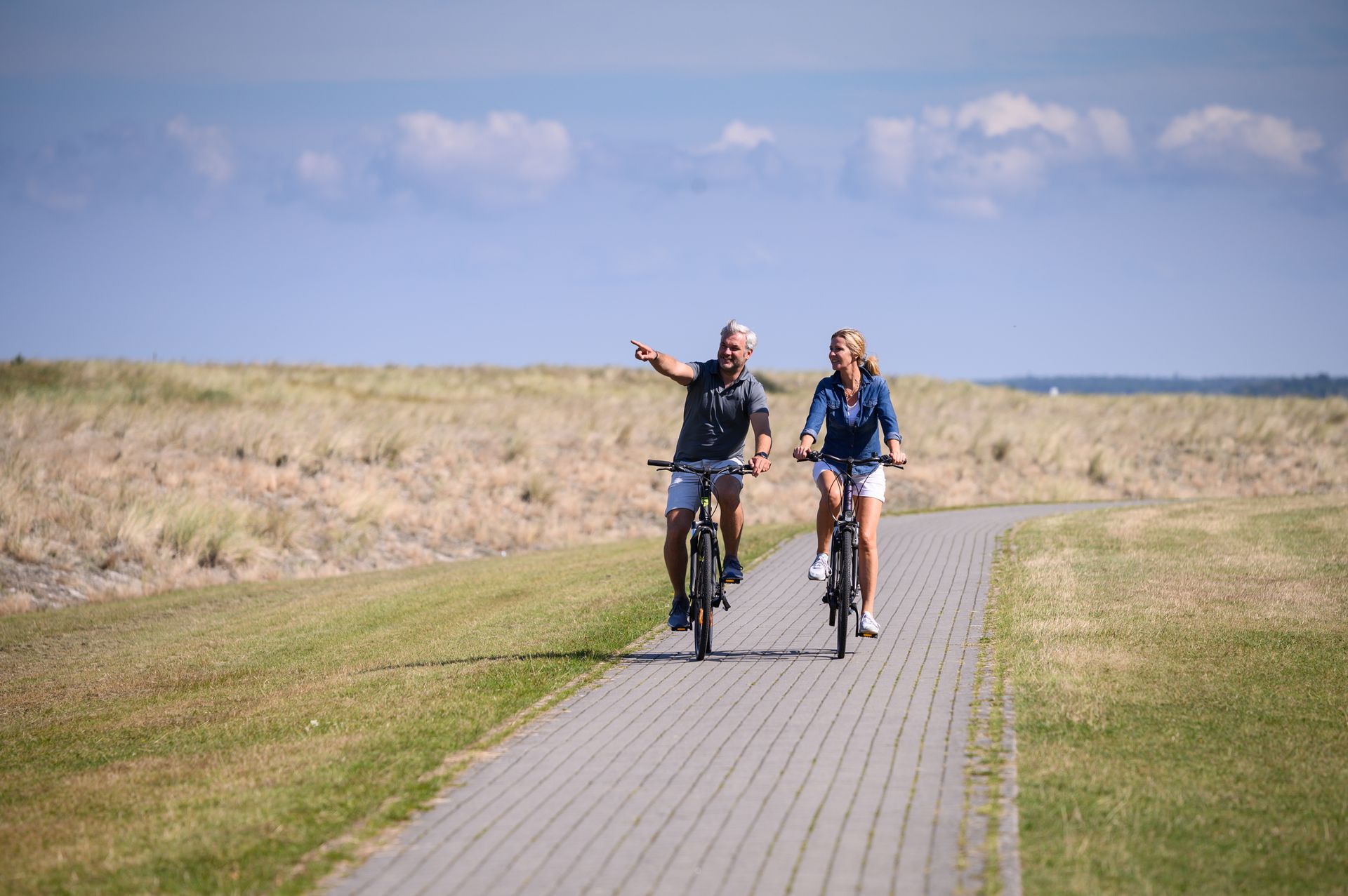 Zwei Radfahrer fahren auf einem asphaltierten Weg neben hohem Gras unter blauem Himmel; einer zeigt mit dem Finger.