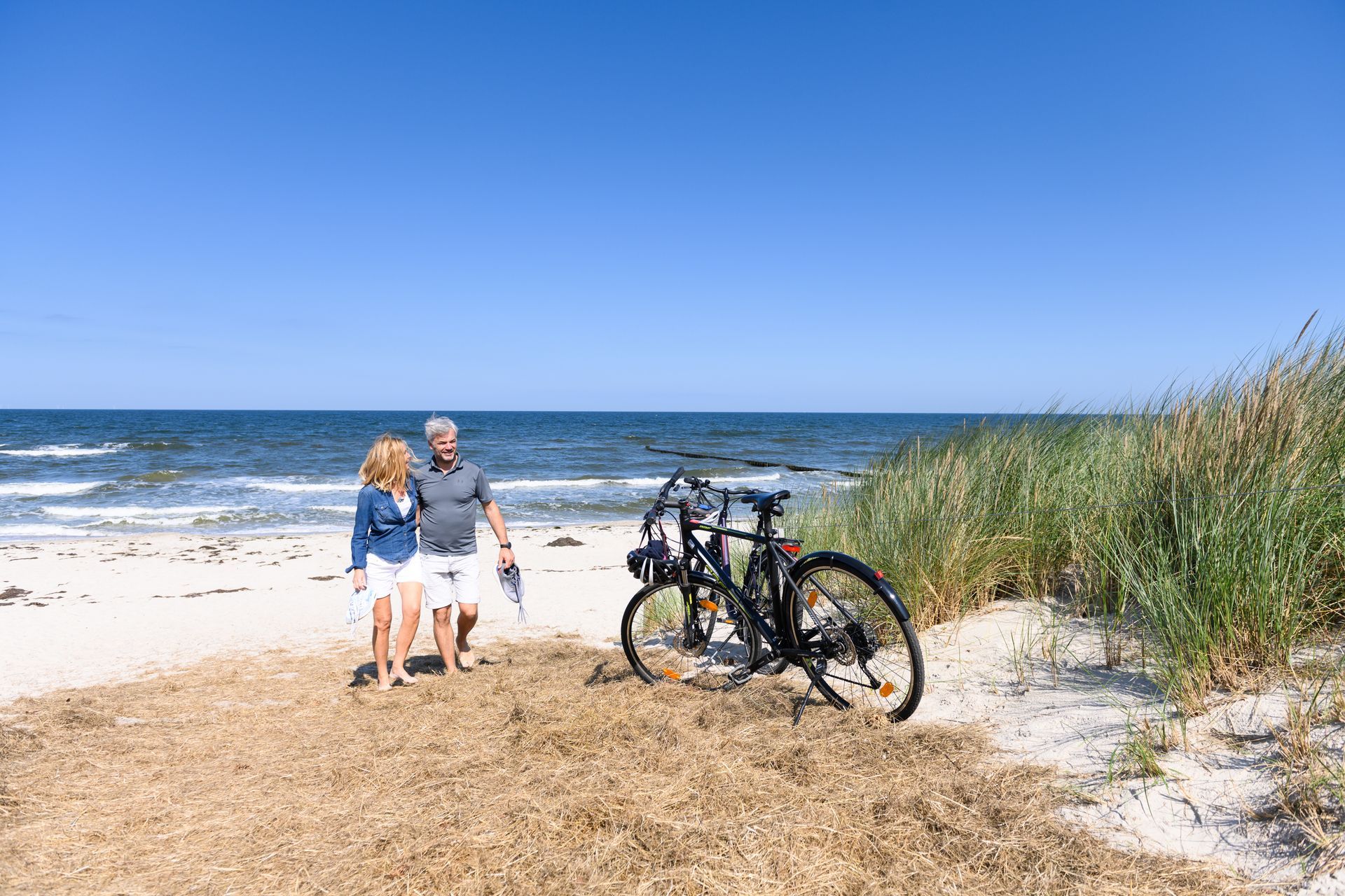 Ein Paar spaziert am Strand entlang, neben einem Fahrrad, blauer Himmel, Meereswellen.