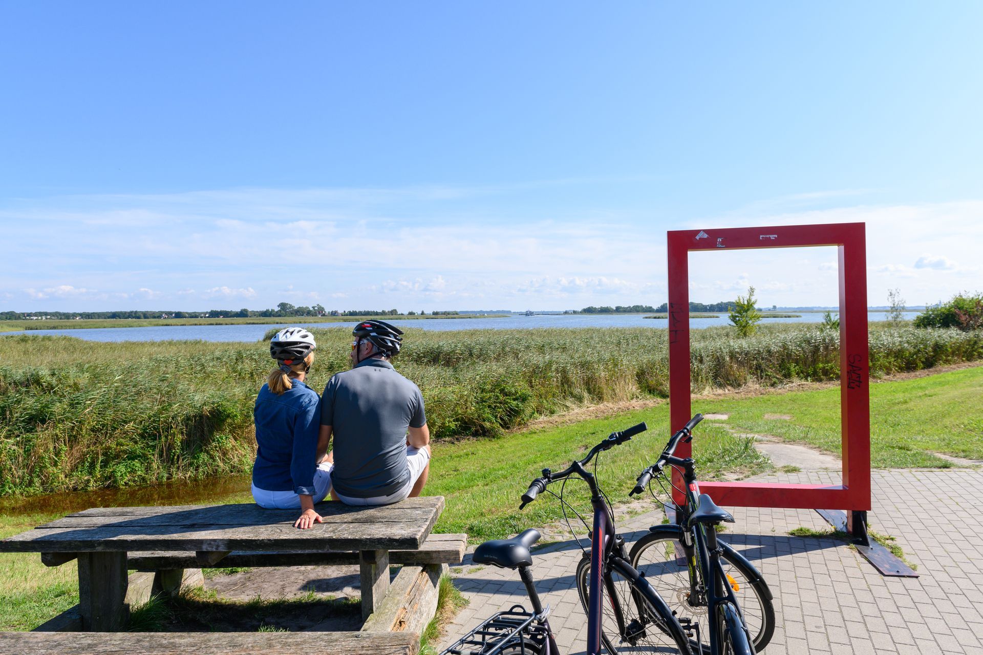 Ein Paar sitzt auf einer Bank und blickt auf ein Gewässer; im Vordergrund sind Fahrräder und ein roter Rahmen zu sehen.