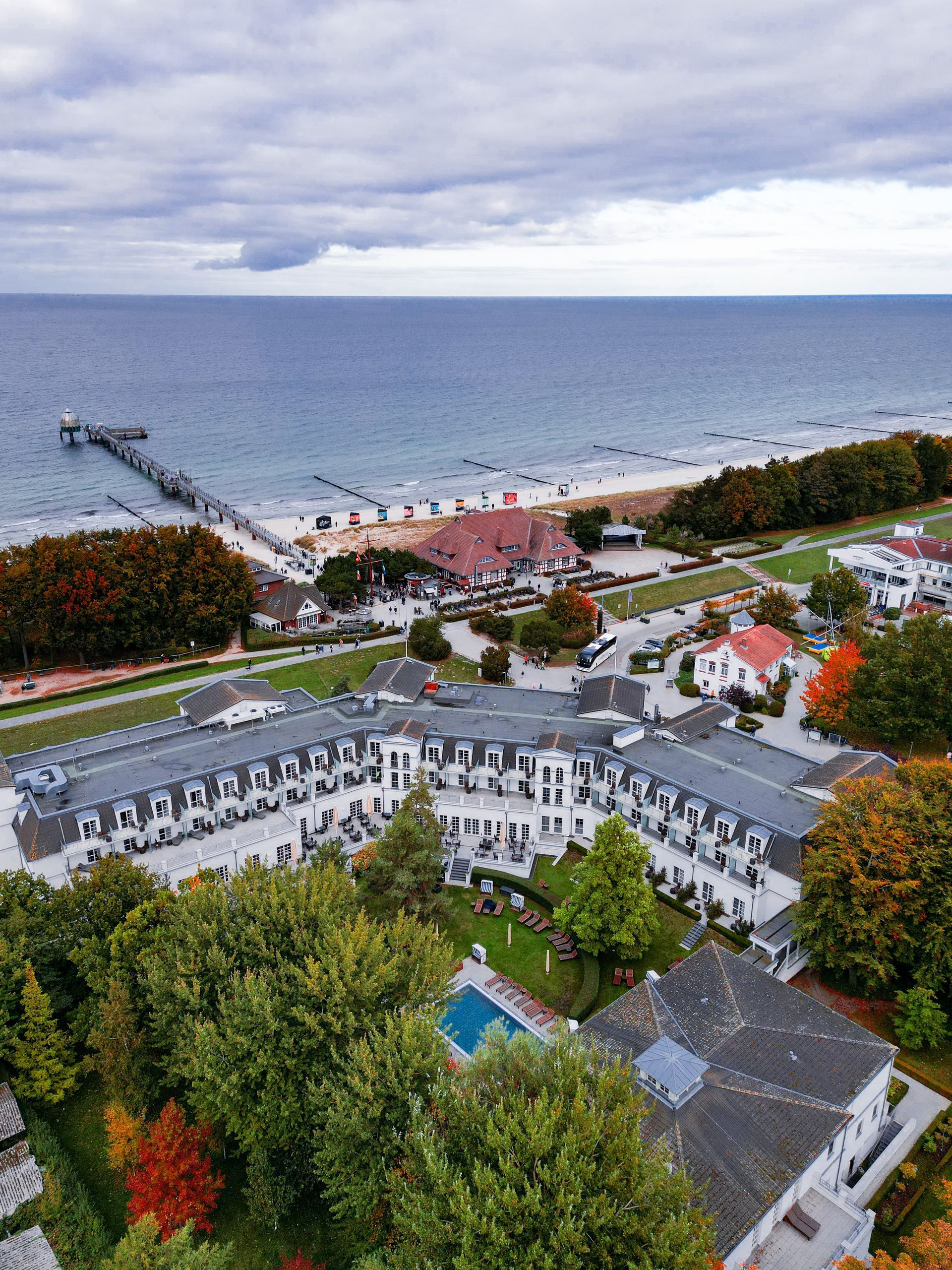 Luftaufnahme eines Küstenhotels mit Pier, Meer und herbstlichen Bäumen unter bewölktem Himmel.