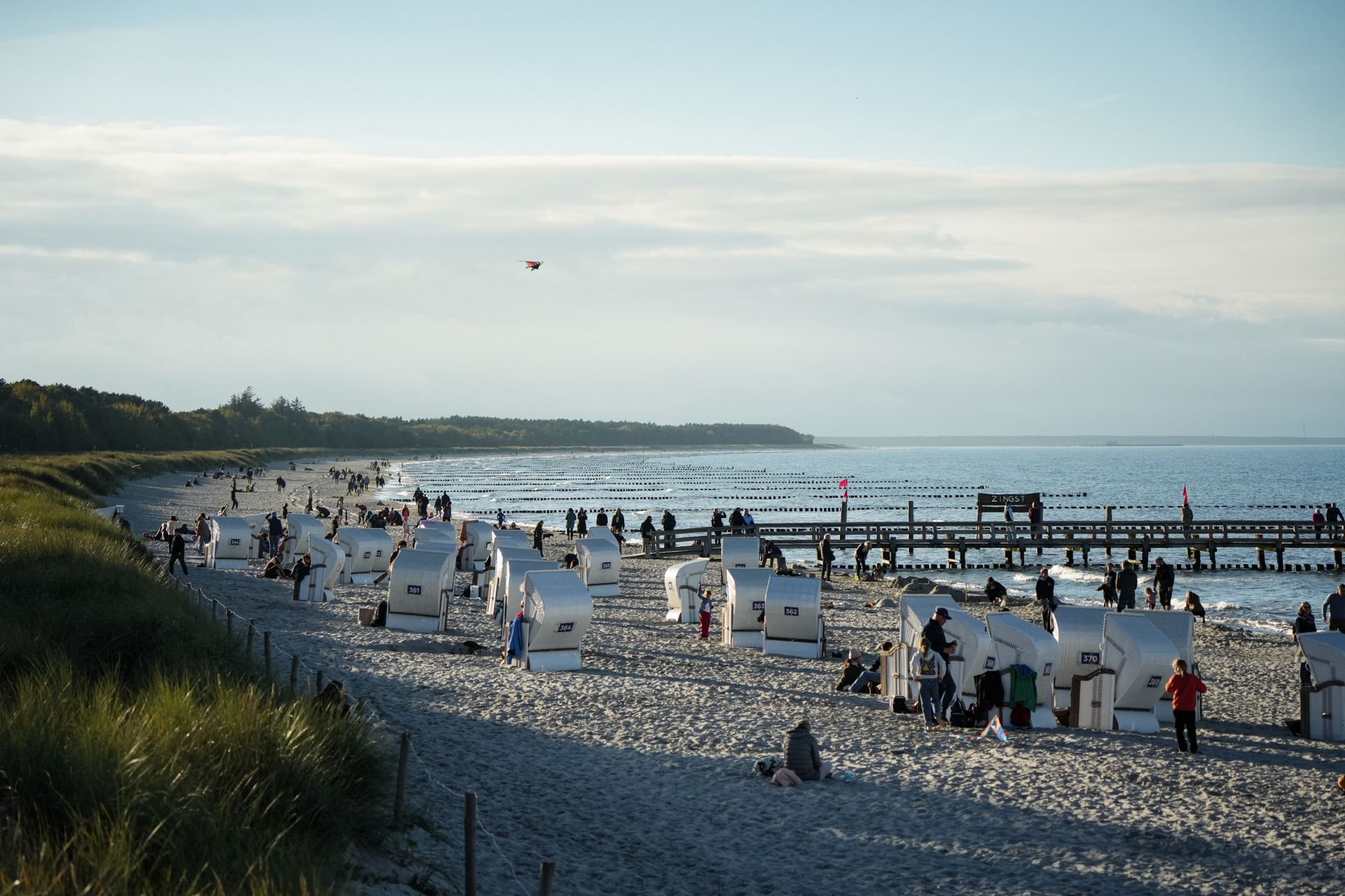 Strandszene mit weißen Strandstühlen, Menschen und einer Seebrücke unter einem teilweise bewölkten Himmel.