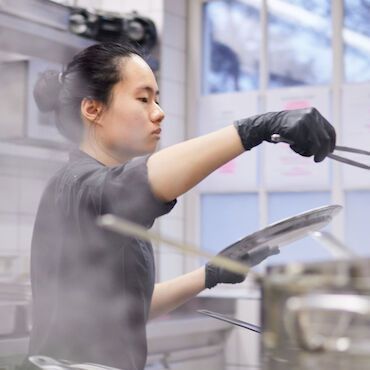 Stellenangebote Köchin mit schwarzen Handschuhen hält in der Küche eine Zange und einen Teller, Dampf steigt auf.