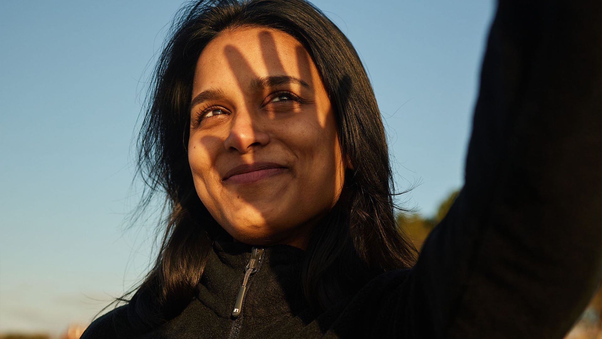 Frau schützt ihre Augen mit der Hand vor der Sonne und lächelt. Hintergrund: blauer Himmel.