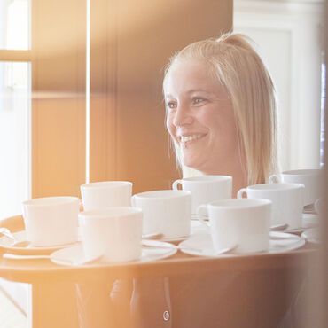 Die Crew vom Strandhotel Zingst Frau hält Tablett mit weißen Teetassen, lächelnd. Heller Raum mit sonniger Beleuchtung.