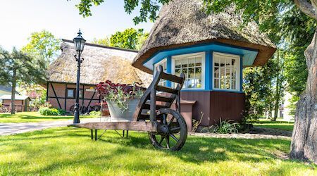 Eine Schubkarre mit Blumen steht neben einem blau umrandeten, strohgedeckten Pavillon.