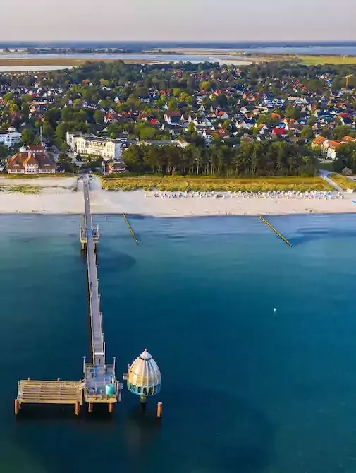 Ein Pier, der in türkisfarbenes Wasser hineinragt, im Hintergrund ein Strand und eine Stadt.