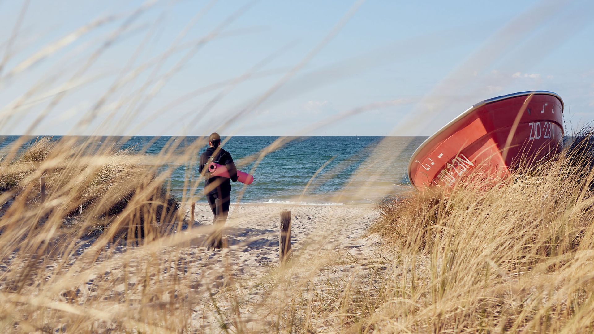 Person, die auf einem Sandweg in Richtung Meer geht, mit Blick auf ein rotes Boot und hohes Gras.