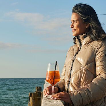 Alle Informationen auf einen Blick Frau im beigen Mantel hält ein Aperol in der Hand und blickt auf die Ostsee.