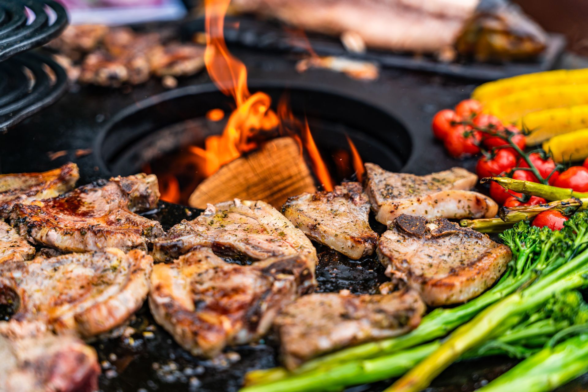 Gegrilltes Fleisch, Mais, Tomaten und Gemüse auf einem Holzgrill mit Flammen.