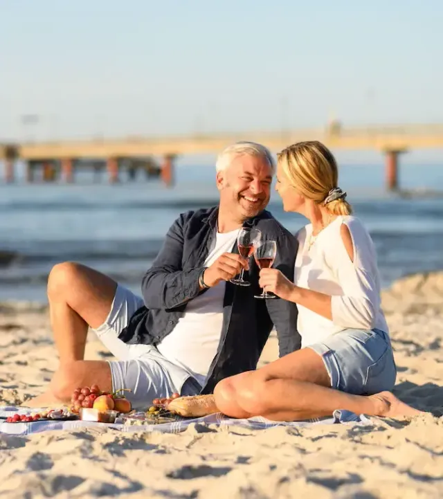 Ein Paar stößt am Strand mit Rotwein an; im Hintergrund sind ein Picknickkorb und eine Seebrücke zu sehen.