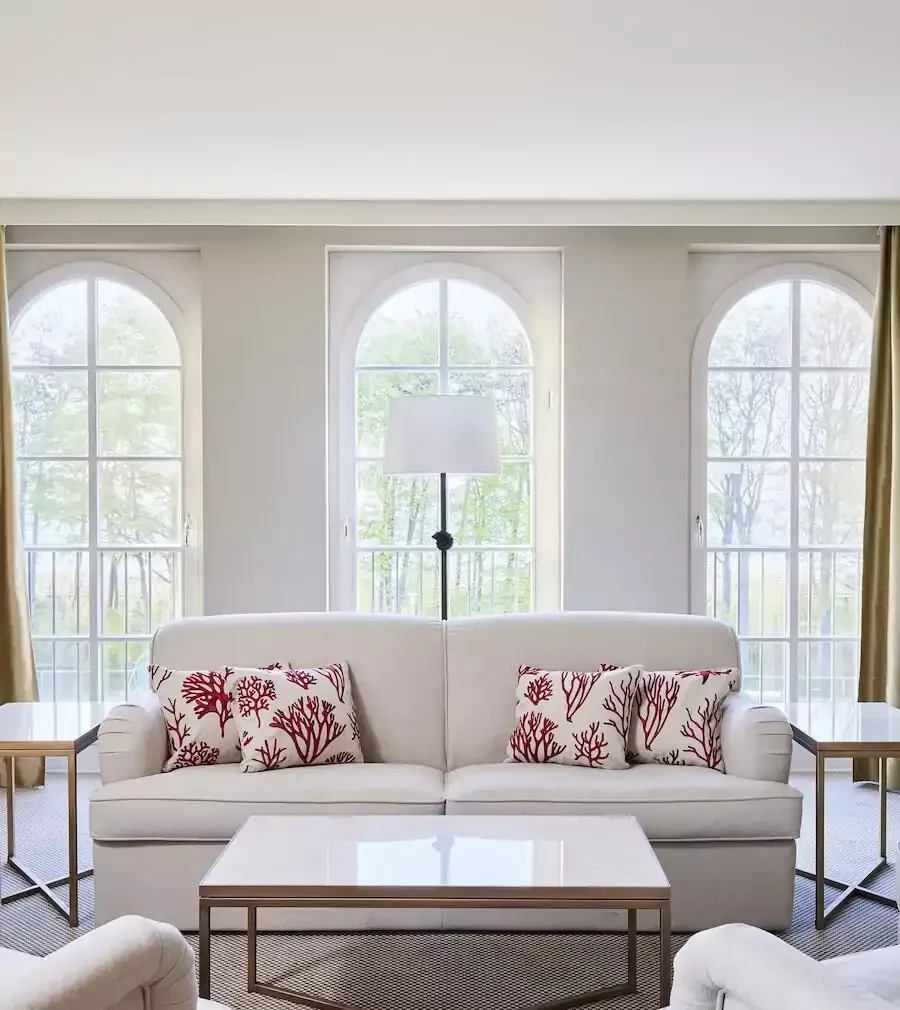 Wohnzimmer mit weißem Sofa, korallenfarbenen Kissen, Bogenfenstern und einem quadratischen Couchtisch.