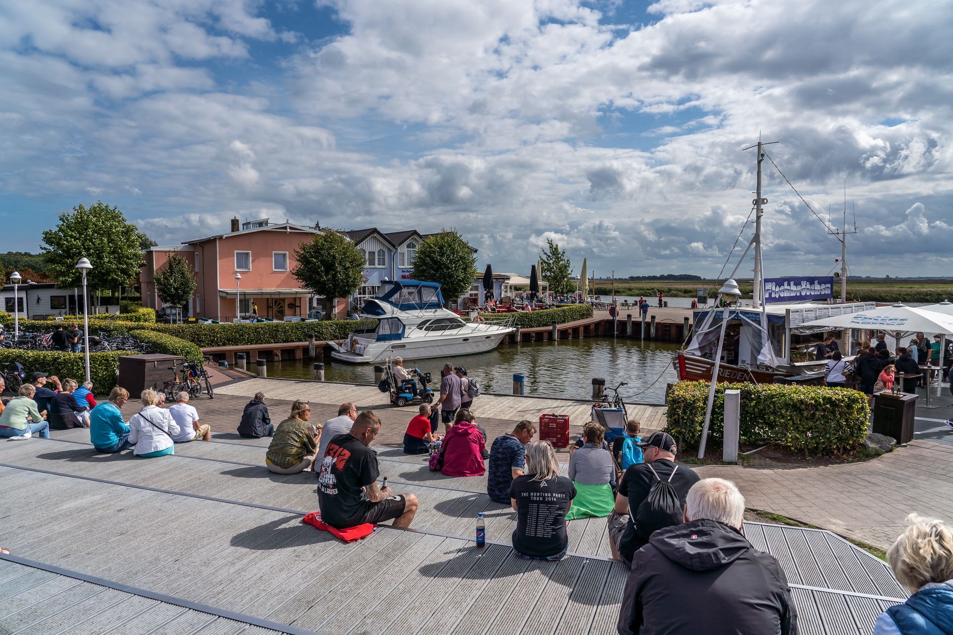 Menschen sitzen auf einem Pier und beobachten ein Geschehen in der Nähe eines Hafens mit Booten.