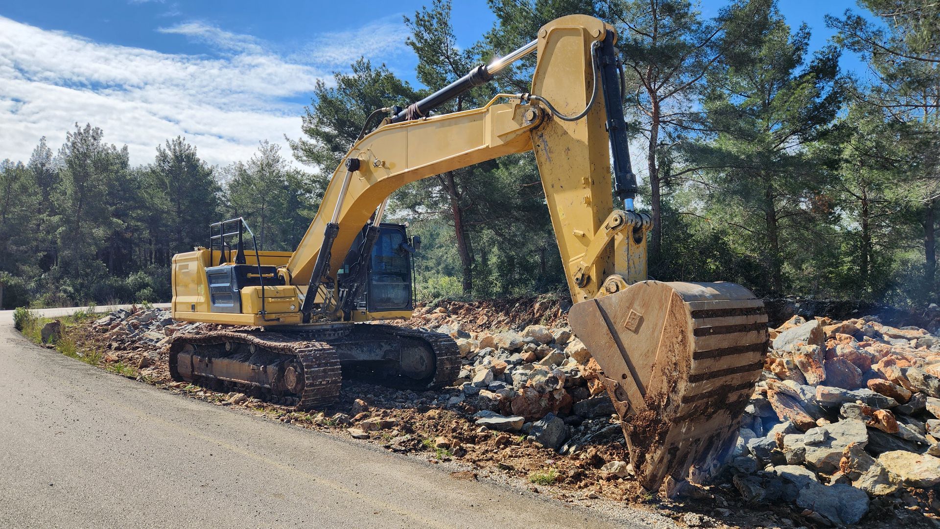 Une pelleteuse jaune sur le bord d'une route de gravier, à côté d'un amas de pierres et d'arbres..