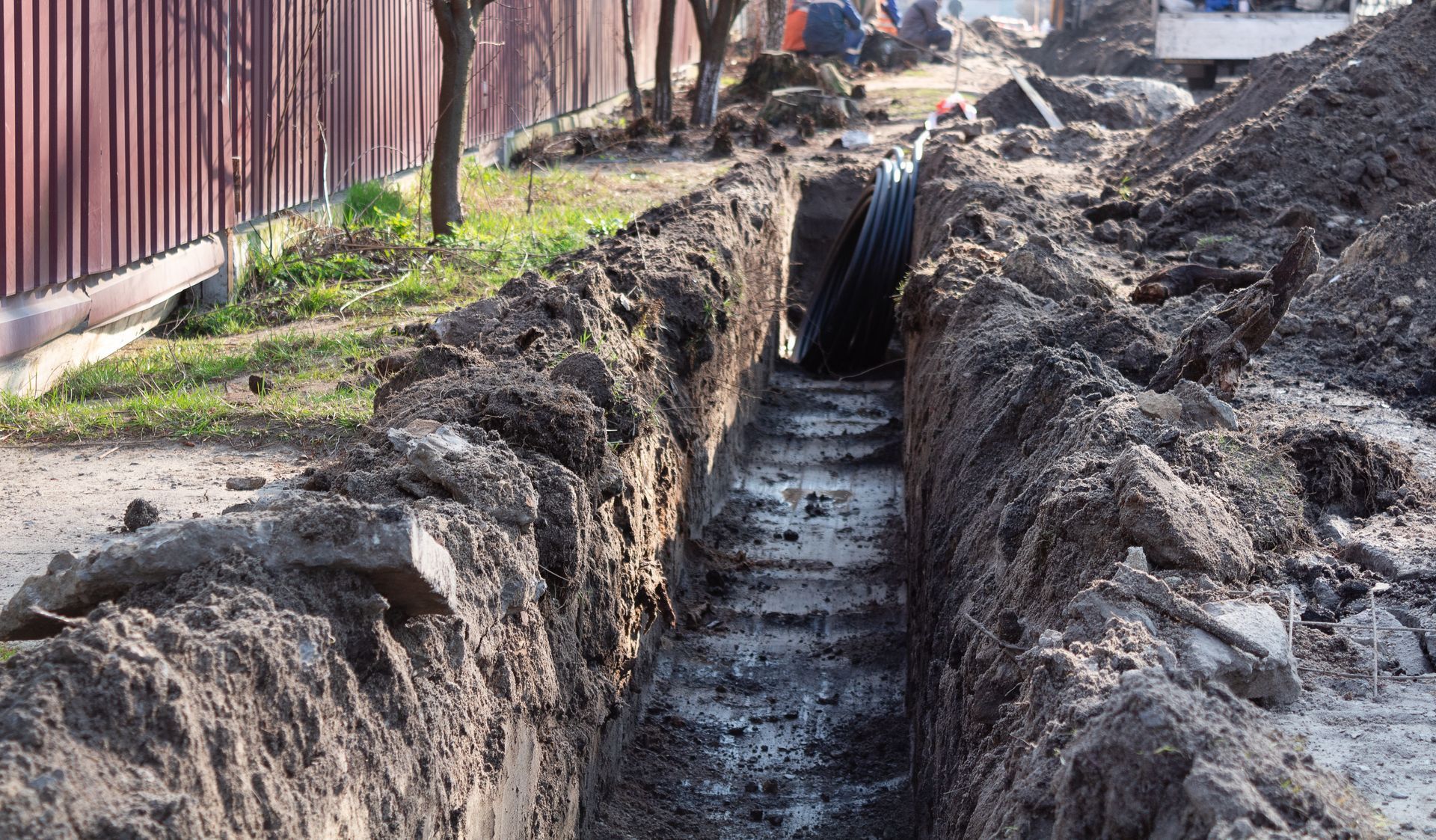 Une profonde tranchée a été creusée le long d'une rue, à côté d'une clôture en bois et de tas de terre sur un chantier.
