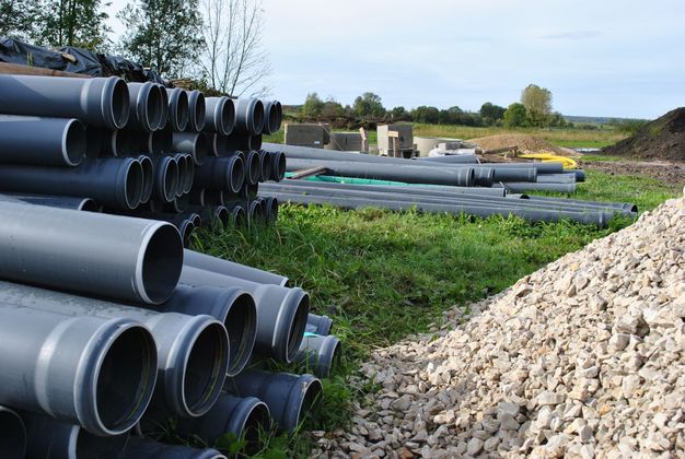 Des piles de gros tuyaux de drainage gris jouxtent un tas de gravier sur un chantier de construction extérieur.