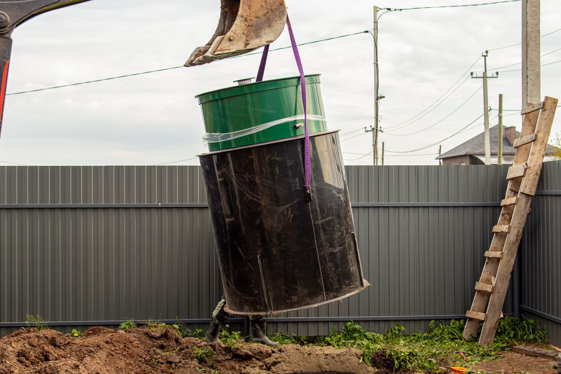 Une excavatrice soulève un baril noir à couvercle vert à côté d'une clôture grise et d'un tas de terre.
