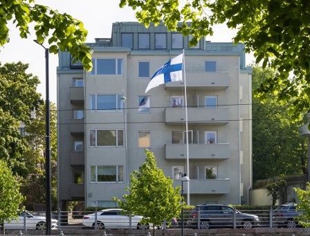 Harmaa rakennus, jonka alla liehuu Suomen lippu; puita ja autoja etualalla.