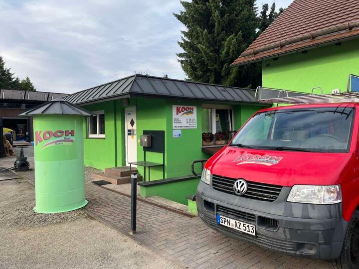 Grünes Gebäude mit schwarzem Dach und davor geparktem roten Lieferwagen; grünes Silo.