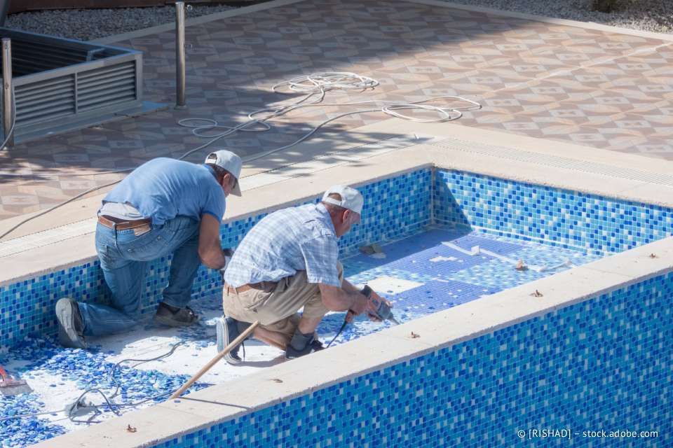 Zwei Männer verlegen Fliesen in einem blau gefliesten Pool und arbeiten im Inneren.